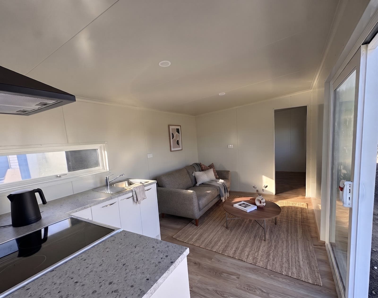 8×3 tiny home open-plan kitchen and living area with induction hob, stone benchtop, white cabinetry and grey sofa