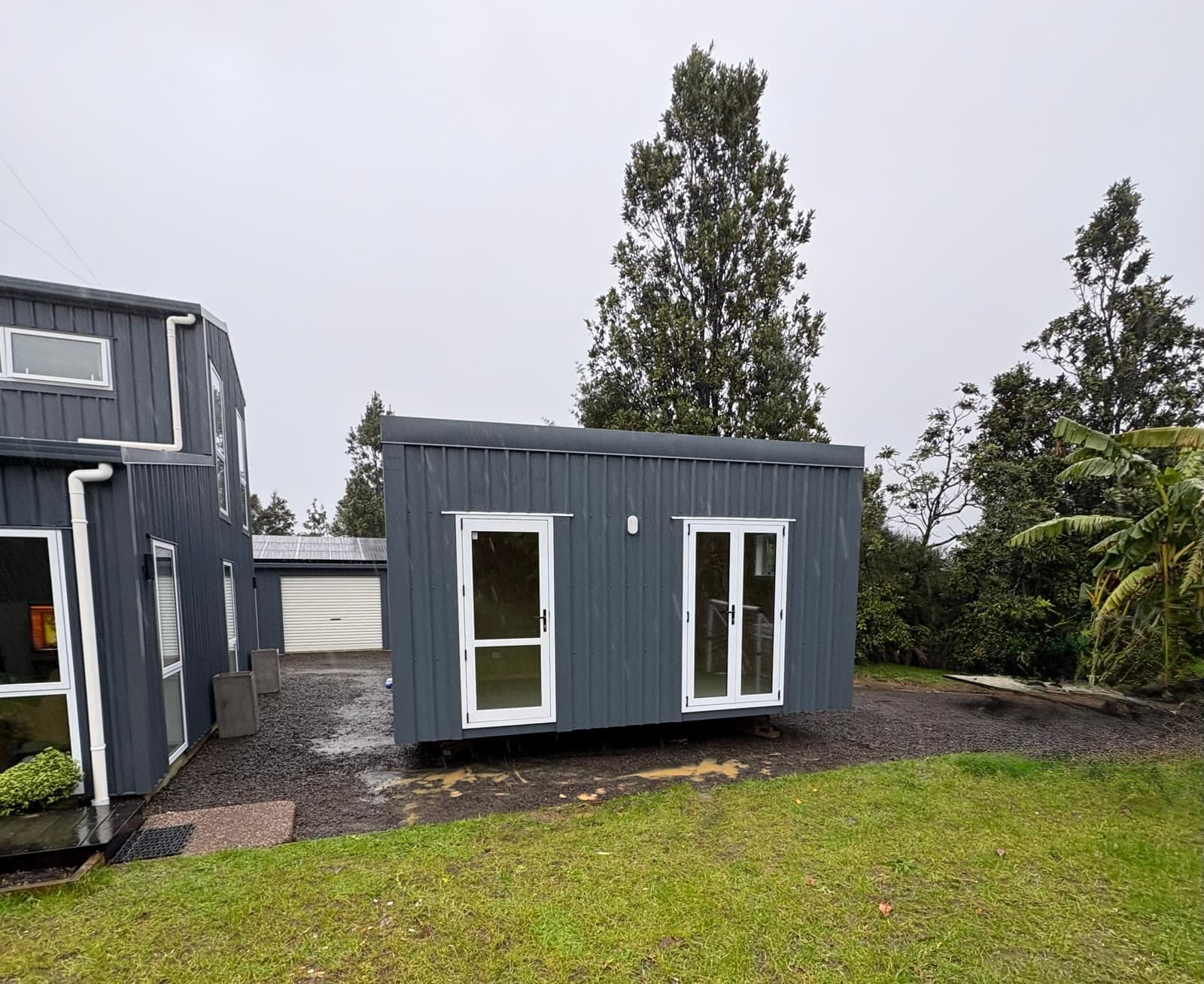 Charcoal-grey NZ-made tiny home with white-trimmed double doors, landed on a gravel pad beside a larger home on a rural section
